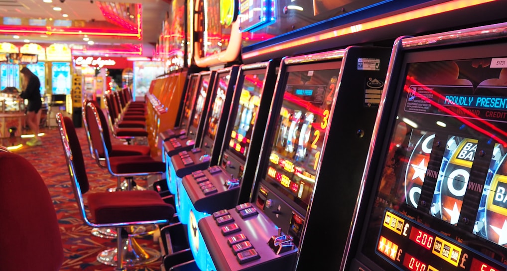 A brightly lit casino interior featuring a row of colorful slot machines. The neon lights create a vibrant atmosphere, with patterned carpeting adding to the decor. Stools are aligned neatly in front of each machine.