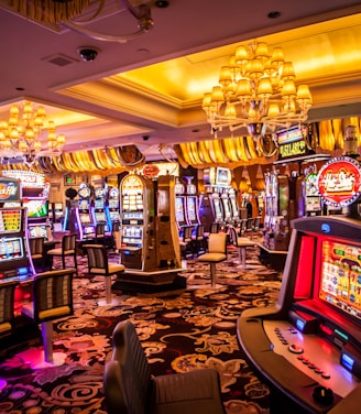 A vibrant casino interior filled with rows of colorful slot machines, each illuminated with bright lights and displays. The ceiling is adorned with elegant chandeliers and golden drapes, creating a luxurious atmosphere. The patterned carpet adds to the elaborate design of the space.