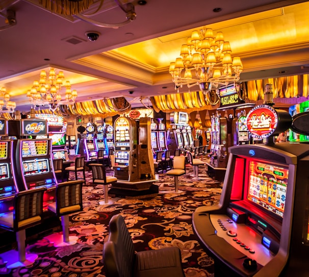 A vibrant casino interior filled with rows of colorful slot machines, each illuminated with bright lights and displays. The ceiling is adorned with elegant chandeliers and golden drapes, creating a luxurious atmosphere. The patterned carpet adds to the elaborate design of the space.