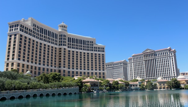 A large luxury hotel and casino building with multiple stories, featuring a light facade with arched windows. The structure has a prominent central tower with a dome-like top. It is surrounded by landscaping with trees and a body of water in the foreground.