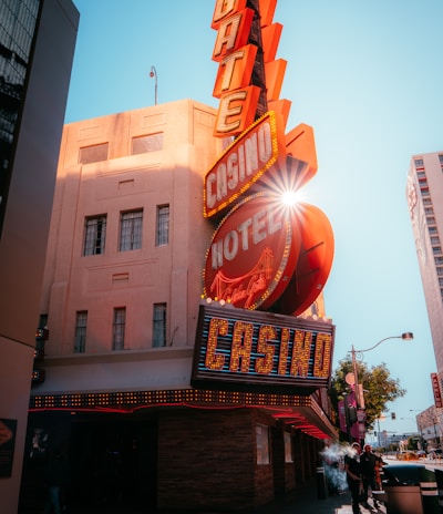 A vibrant city scene featuring a tall, illuminated casino sign with retro-style neon lights. The sign dominates the building facade with the words 'CASINO' and 'HOTEL'. Sunlight peeks through, creating a dramatic lens flare effect. The street is lively, with pedestrians casually walking and a hint of urban hustle.